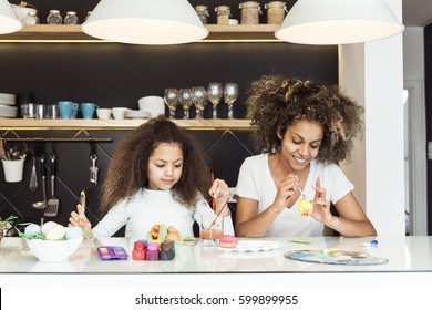 Beautiful African American woman and her daughter coloring Easter eggs in the kitchen 