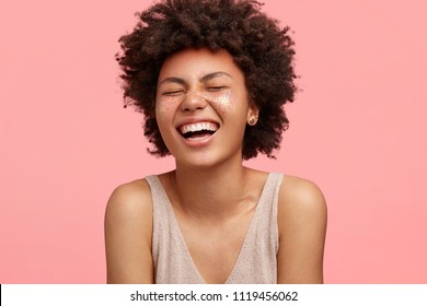 Beautiful African American woman laughs at something funny, has sparkles on cheeks, shows bare shoulders, closes eyes with pleasure, isolated over pink background. People and make up concept