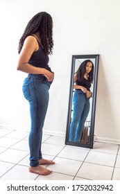 Beautiful African American Woman And Mother Stands In Front Of A Mirror And Attempts To Try To Close Her Tight Fitting Blue Jeans Pants After Postpartum Diet And Weight Loss Exercise.