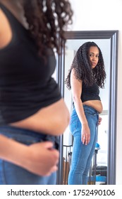 Beautiful African American Woman And Mother Stands In Front Of A Mirror And Attempts To Try To Close Her Tight Fitting Blue Jeans Pants After Postpartum Diet And Weight Loss Exercise.