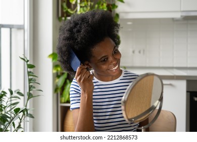 Beautiful African American Woman Takes Care Of Hair Looks In Mirror With Plastic Comb And Smiles Broadly. Black Lady In Striped T-shirt Doing Hairstyle For Party Sits In Kitchen With Home Plants