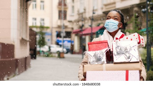 Beautiful African American Woman Walking In Decorated City With Many Christmas Gift Boxes After Xmas Shopping. Portrait Of Joyful Grateful Female In Mask Holds Holiday Presents. Surprise Gifts Concept