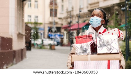 Beautiful African American woman walking in decorated city with many christmas gift boxes after xmas shopping. Portrait of joyful grateful female in mask holds holiday presents. Surprise gifts concept