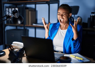 Beautiful african american woman working at the office at night crazy and mad shouting and yelling with aggressive expression and arms raised. frustration concept.  - Powered by Shutterstock
