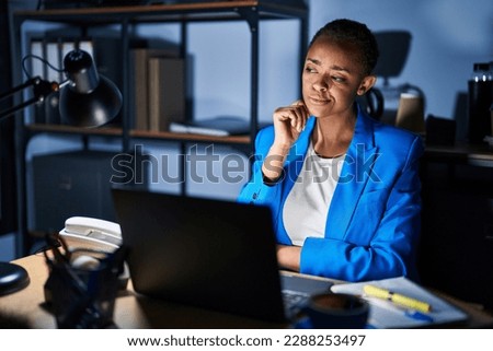 Beautiful african american woman working at the office at night with hand on chin thinking about question, pensive expression. smiling with thoughtful face. doubt concept. 