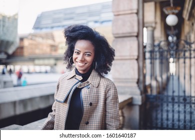 Beautiful african american young woman with afro and large hoop earrings in a stylish coat, smiling. Urban street portrait.