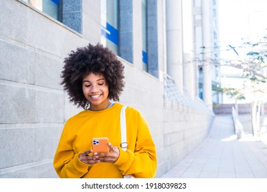 beautiful African or American young woman walking in the street of the city looking at her phone smiling and having fun enjoying