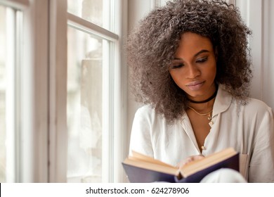 Beautiful african american young woman sitting by the window and reading a book.