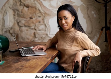 Beautiful African Business Woman Is Reading Emails On A Laptop While Sitting In A Cafe. A Beauty Blogger Is Watching The New Fashion Show On-line On A Portable Computer Connected To The Wifi.