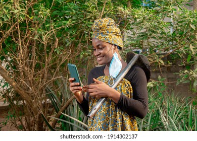 
A Beautiful African Female Farmer With Nose Mask, Checking Her Smartphone With A Hoe Hanging From Her Shoulder In Her Pineapple Farm