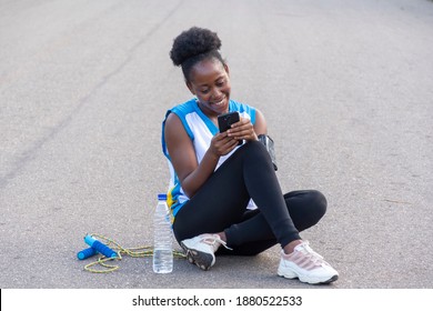 Beautiful African Lady, After An Exercise, Sitting Down On The Floor, Smiling And Using Her Phone