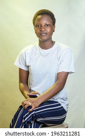 A Beautiful African Lady Sitting Down With Legs Crossed And Wearing A White Shirt With A Phone In Her Hand