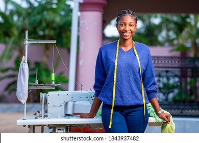 Beautiful African seamstress standing in front of sewing machine with measuring tape around neck,cheerfully looking at camera-concept on millennial black female entrepreneurship 
