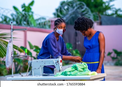 Beautiful African seamstress wearing face mask and measuring a client in front of sewing machine in outdoor studio-black tailor at work in covid-19 pandemic-concept on business and style and fashion. - Powered by Shutterstock