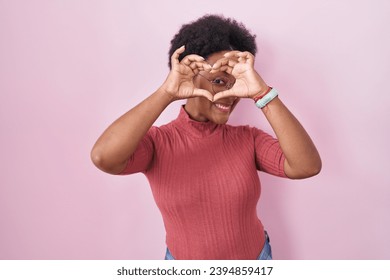 Beautiful african woman with curly hair standing over pink background doing heart shape with hand and fingers smiling looking through sign  - Powered by Shutterstock