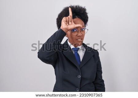 Beautiful african woman with curly hair wearing business jacket and glasses making fun of people with fingers on forehead doing loser gesture mocking and insulting. 
