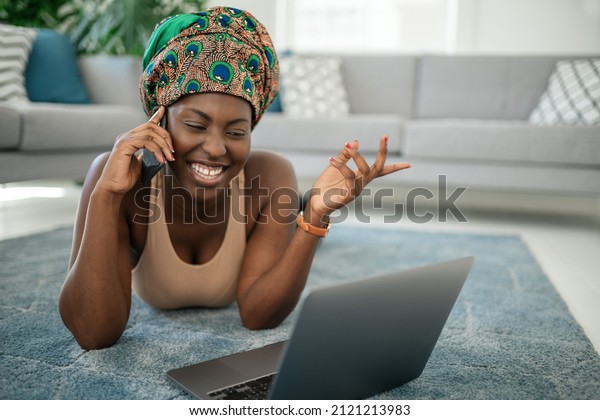 Beautiful African woman at home, smiling and\
laughing, using mobile smart phone, wearing traditional headscarf,\
surprised and happy