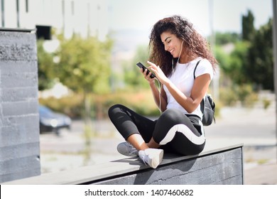 Beautiful African Woman Listening To Music With Earphones And Smart Phone Outdoors. Arab Girl In Sport Clothes With Curly Hairstyle In Urban Background.