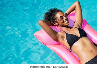 Beautiful african woman lying on pink mattress in outdoor swimming pool. Young black woman sunbathing in swimming pool with copy space. Happy girl in glamour bikini relaxing in pool with eyeglasses. - Powered by Shutterstock