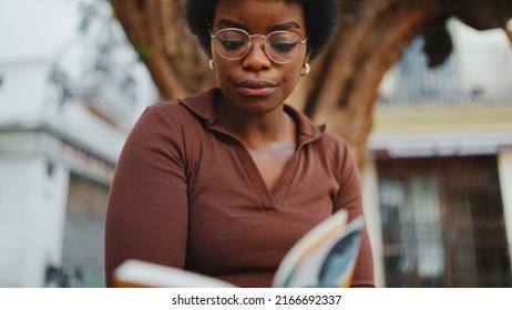 Beautiful African Woman Sitting With Book On The Street, Wearing Glasses. Close Up Dark-skinned Girl Reading Book Outdoors