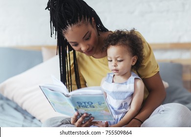 Beautiful African woman sitting on bed and reading a book to her cute baby daughter.