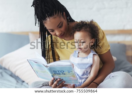 Beautiful African woman sitting on bed and reading a book to her cute baby daughter.