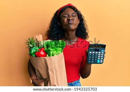 Beautiful african young woman holding groceries and calculator puffing cheeks with funny face. mouth inflated with air, catching air. 