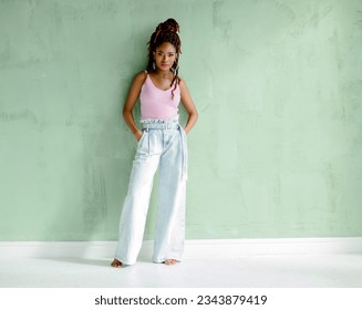 Beautiful African young woman looking at the camera, standing barefoot with her hands in pockets, isolated on a light green wall with copy space. Full-length fashion portrait of an attractive model. - Powered by Shutterstock