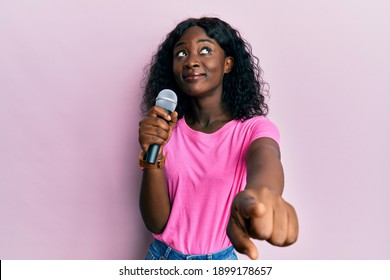 Beautiful African Young Woman Singing Song Using Microphone Pointing To Camera Smiling Looking To The Side And Staring Away Thinking. 