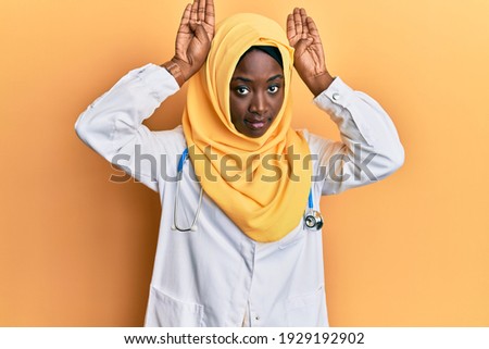 Beautiful african young woman wearing doctor uniform and hijab doing bunny ears gesture with hands palms looking cynical and skeptical. easter rabbit concept. 
