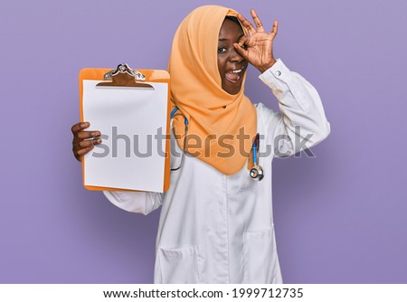 Beautiful african young woman wearing doctor stethoscope holding clipboard smiling happy doing ok sign with hand on eye looking through fingers 