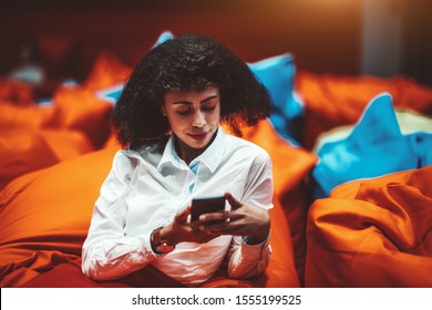 Beautiful African-American businesswoman is relaxing on an orange cushion in a lounge area of her office and using a smartphone; a biracial woman entrepreneur with a cellphone in a chillout zone