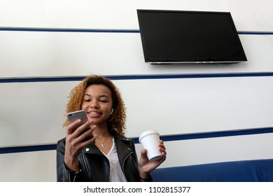 A Beautiful African-American Girl In A Black Jacket With Airpods In Her Ear Smiles And Holds A White Glass And Looks Into The Phone. TV On A White Wall With Blue Stripes Of The Airport Waiting Room.