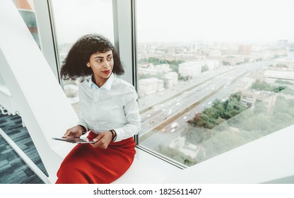 A Beautiful African-American Woman Entrepreneur In A White Blouse And Red Skirt Is Holding A Digital Tablet Pc While Sitting Indoors On The Bench On The Top Floor Of A Business Office Skyscraper