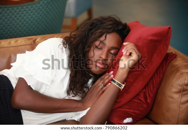 Beautiful African-American woman\
keeping head on red pillows while taking nap on comfortable sofa.\
