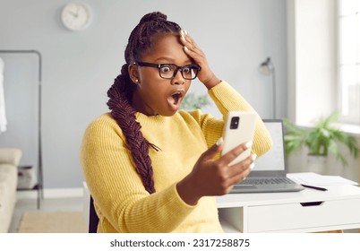 Beautiful africanamerican woman looks at the phone in horror with her mouth and eyes wide open. Stunned dark-skinned girl dressed in a yellow sweatshirt holds her head while looking at her phone.