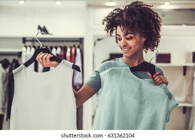 Beautiful Afro American girl is choosing a dress and smiling while doing shopping in female boutique