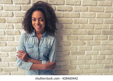 Beautiful Afro American girl is looking at camera and smiling while standing with crossed arms against white brick wall - Powered by Shutterstock