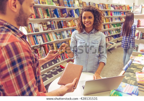 Beautiful afro american girl is\
smiling while buying book at the bookshop using a credit\
card