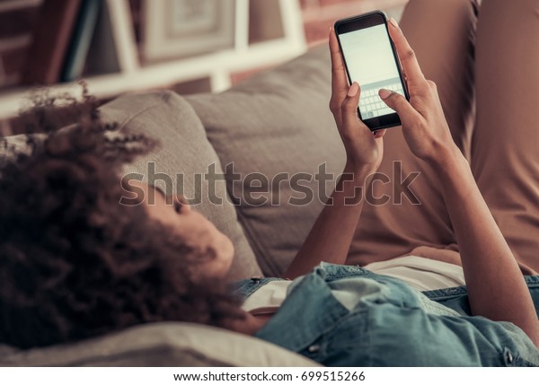 美しいアフロ系アメリカ人の女の子は 家のソファに寝そべりながら スマートフォンを使って微笑んでいる の写真素材 今すぐ編集