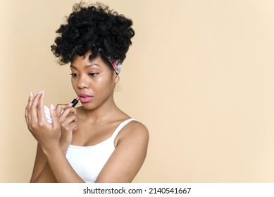 Beautiful afro american woman applying lipstick on her lips and looking in the mirror on a beige background. opy space
