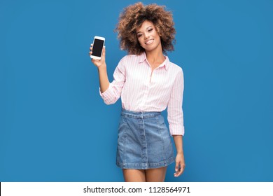 Beautiful afro woman showing mobile phone and smiling, standing on blue background. Girl wearing jeans short skirt and elegant shirt in white and pink stripes. - Powered by Shutterstock