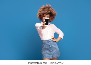 Beautiful afro woman showing mobile phone and smiling, standing on blue background. Girl wearing jeans short skirt and elegant shirt in white and pink stripes. - Powered by Shutterstock