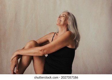 Beautiful Aging Woman Celebrating Her Natural Body In A Studio. Mature Woman In Black Underwear Smiling Cheerfully While Sitting Alone Against A Studio Background.