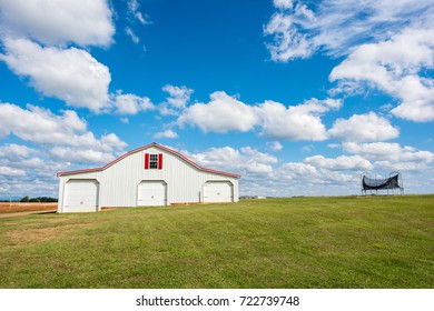 Beautiful all american red and white barn in a grassy field in southern america with a bright, blue sky and a trampoline beyond.