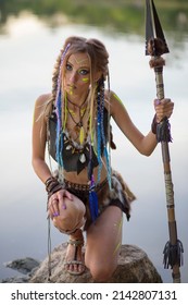 Beautiful Amazon Woman In A Fur Medieval Costume, In War Paint, In The Jungle With A Spear Against The Background Of The River.