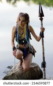 Beautiful Amazon Woman In A Fur Medieval Costume, In War Paint, In The Jungle With A Spear Against The Background Of The River.