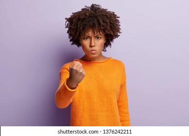 Beautiful annoyed woman raises fist, has frustrated and furious outraged expression, expresses aggression, rage, dressed in orange jumper, isolated over purple background, promises to revenge.