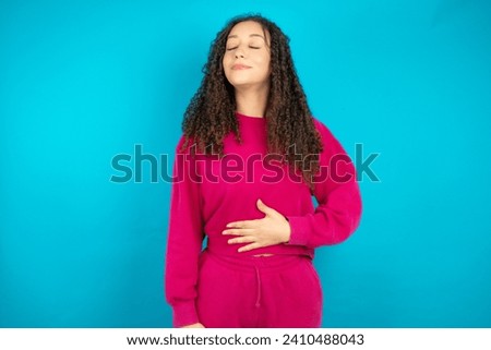 Beautiful Arab teen girl with curly hair wearing pink sweater touches tummy, smiles gently, eating and satisfaction concept.