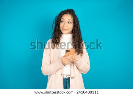 Beautiful arab teen girl with curly hair wearing turtleneck sweater holding in hands mobile phone and looking aside with dreamy look.